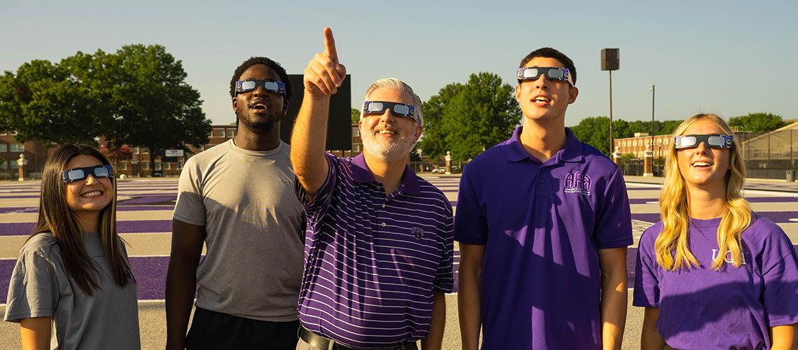 President Houston Davis and several students gaze at the sky through eclipse glasses. The picture is part of UCA's promotional material for the April 2024 eclipse at uca.edu/eclipse.
